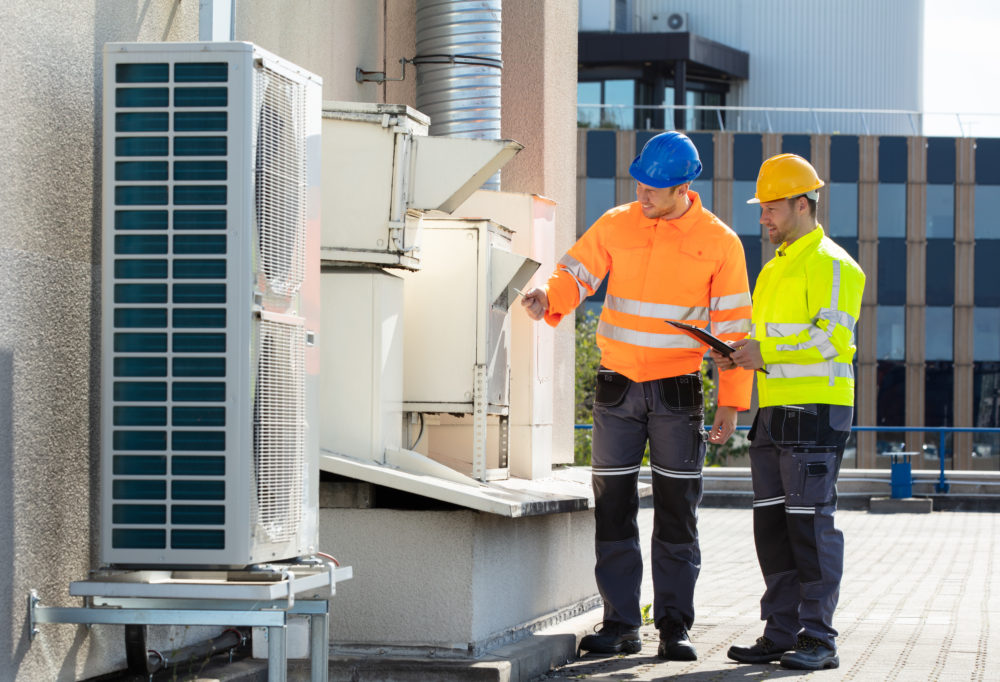 Two Electricians Men Wearing Safety Jackets Checking Air Conditioning Unit On Building Rooftop